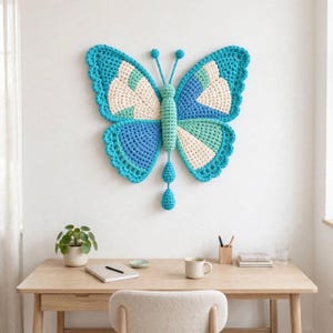 May include: A large, crocheted butterfly wall hanging featuring a color palette of turquoise, light blue, and cream. The butterfly has a central body with two antennae and a dangling tail, all crafted with a textured yarn stitch pattern.