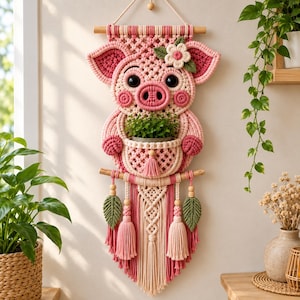 Könnte beinhalten: Ein Makramee-Wandbehang in Schweineform mit einer zentralen Tasche, die eine kleine grüne Pflanze enthält. Das Design besteht aus cremefarbenen und rosafarbenen geknoteten Schnüren, großen schwarzen Augen, einer Blume und Quasten.