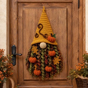 Può includere: Una decorazione da parete a forma di gnomo all'uncinetto con un alto cappello giallo senape, decorazioni a spirale marroni e una piccola zucca arancione. La barba è composta da fili di lana ricci in colori autunnali con zucche e foglie.