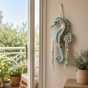 Puede incluir: Un tapiz de pared con forma de caballito de mar, elaborado con cuerda de color azul claro y crema mediante técnicas de macramé. El caballito de mar presenta una cuenta oscura como ojo, conchas decorativas a lo largo del lomo y flecos.