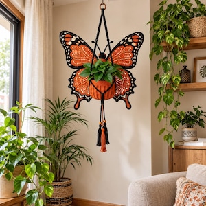May include: A hanging crochet plant holder shaped like a monarch butterfly with orange and black wings. The central orange crochet basket holds a green plant. The structure is suspended by black cords and features decorative black and orange tassels.