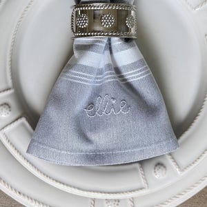 May include: A formal place setting with a white plate, a gray and white striped napkin, and silver silverware. The napkin is held by a silver napkin ring. The plate has a decorative border. The word "Ellie" is embroidered on the napkin.