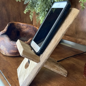 May include: A wooden phone stand with a smartphone in a black case. The stand is made of light-colored wood and has a simple, angled design. The phone is black with a white screen and a black case.