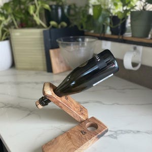 May include: A dark glass wine bottle resting at an angle in a two-piece wooden stand. The stand is crafted from light brown wood with visible grain, featuring the number 45 stamped on the base and the upper support piece holding the bottle neck.