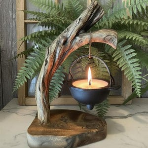 May include: A small black metal cauldron candle holder suspended from a curved piece of natural driftwood. The driftwood is attached to a flat, polished wooden base. A single lit tea light candle sits inside the cauldron, emitting a warm glow.