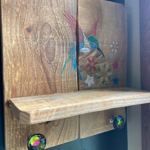 May include: A rustic wooden shelf with a hummingbird design. The shelf is made of light-colored wood and features a painted hummingbird with red, blue, and green accents. It has a small shelf and is adorned with two iridescent crystal knobs.