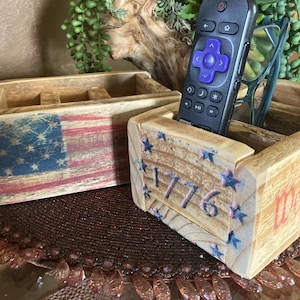 May include: Two wooden storage boxes with patriotic designs. One box features a painted American flag, the other has the number "1776" with blue star accents. A remote control and glasses rest inside one box.