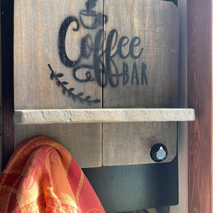 May include: A rustic wooden coffee bar sign with the words "Coffee Bar" stenciled in black. The sign has a small shelf and a decorative knob. A red and orange patterned towel hangs below.