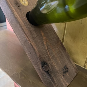 May include: A dark brown stained wooden wine bottle holder angled to support a green glass bottle. The holder features a circular brass-colored metal inlay at the top and the number 45 stamped into the lower front surface of the wood.