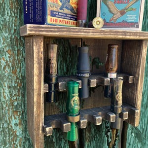 May include: A rustic wooden shelf displaying hunting calls and vintage shotgun shell boxes. The shelf holds various duck calls in different colors and designs. The boxes feature illustrations of ducks and text, including "High Velocity" and "Hi-Power Shot Shells."