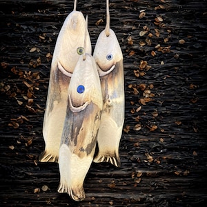 May include: Three wooden fish ornaments hanging by twine. The fish have a natural wood color with darker brown accents and blue, green, and gray eyes. The fish are carved with fins and a smiling mouth. The background is a dark, textured surface.