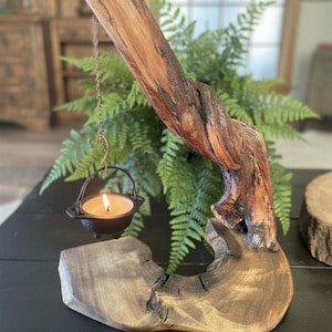 May include: A natural wood candle holder featuring a curved, textured branch extending from a flat, circular wooden base. A small, black, three-legged metal cauldron hangs from the branch by a thin chain, containing a lit tea light candle.