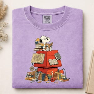 May include: A light purple crewneck t-shirt featuring a graphic of Snoopy wearing glasses and reading a book while sitting atop a red doghouse. The doghouse is surrounded by stacks of books, a globe, a quill, and a desk lamp. The label reads COMFORT COLORS.