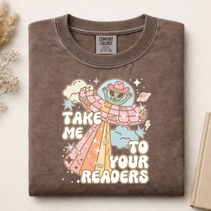 May include: A brown crewneck t-shirt featuring a graphic of a green alien wearing a pink cowboy hat inside a flying saucer. The text reads "TAKE ME TO YOUR READERS" in a bold, retro font, surrounded by stars, clouds, and small book icons.