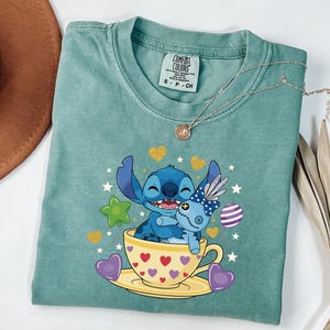 May include: A teal Comfort Colors t-shirt with a graphic of Stitch from Lilo & Stitch hugging a doll in a teacup. The teacup is yellow with red hearts. The shirt has a small tag and a silver necklace.