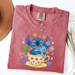 May include: A red-pink t-shirt with a graphic of Stitch from Lilo & Stitch, hugging a doll in a teacup. The teacup is yellow with red hearts. A silver necklace with a pendant is draped on the shirt.