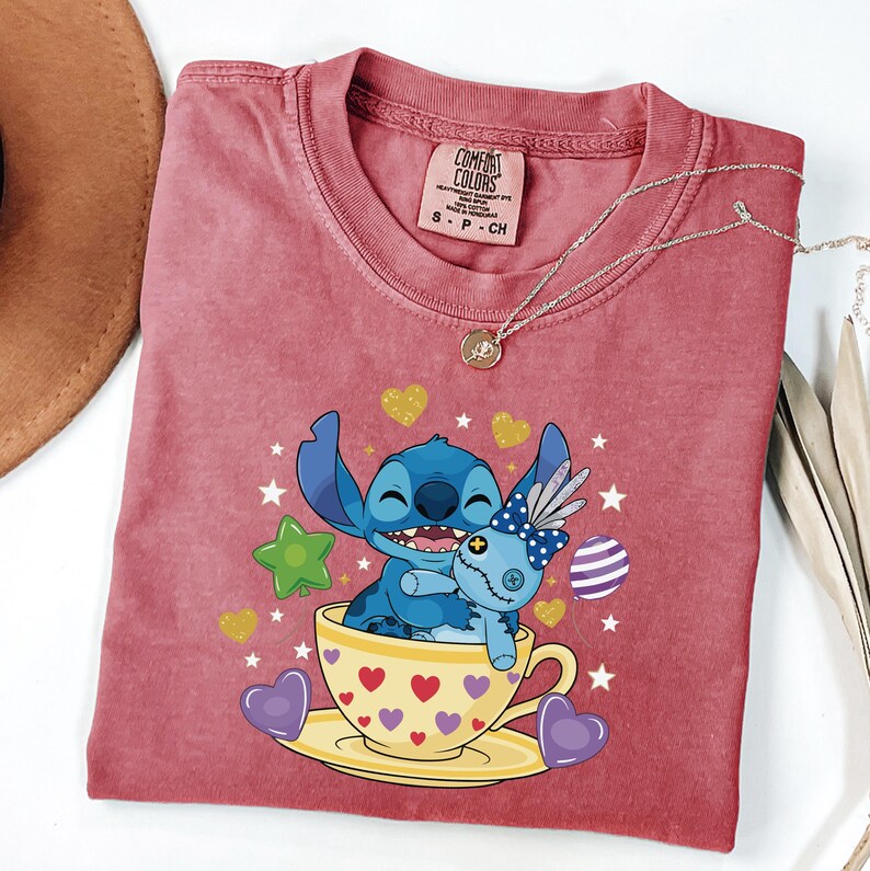 May include: A red-pink t-shirt with a graphic of Stitch from Lilo & Stitch, hugging a doll in a teacup. The teacup is yellow with red hearts. A silver necklace with a pendant is draped on the shirt.