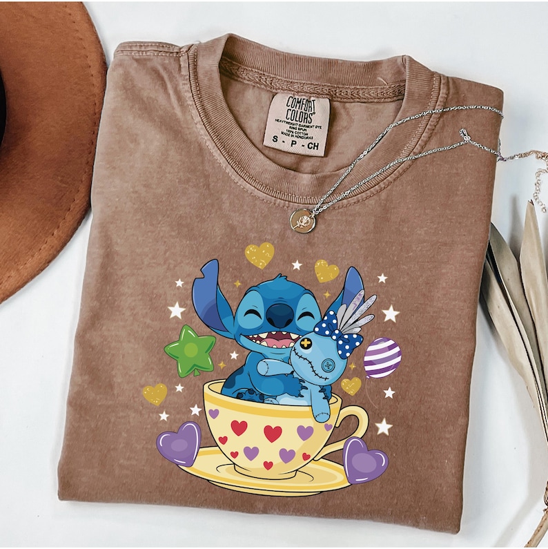 May include: Brown Comfort Colors t-shirt with a cartoon image of Stitch and a doll in a teacup. The teacup is yellow with red hearts. The shirt has a crew neck and short sleeves. A silver necklace is also visible.