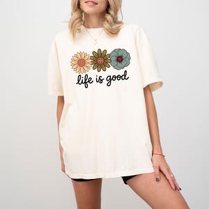 May include: Cream-colored t-shirt featuring three floral designs in shades of orange, green, and teal. The phrase "life is good" is printed in a black script font below the flowers. The shirt has a relaxed fit.