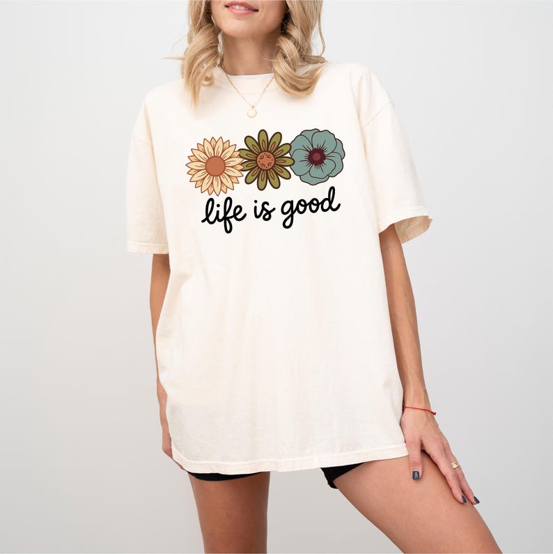 May include: Cream-colored t-shirt featuring three floral designs in shades of orange, green, and teal. The phrase "life is good" is printed in a black script font below the flowers. The shirt has a relaxed fit.