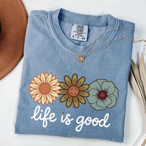 May include: A light blue t-shirt with three floral designs in yellow, green, and blue, and the words "life is good" in white script. A gold necklace with a pendant rests on the shirt. The shirt is folded on a white surface.
