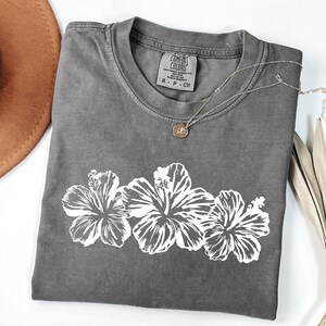 May include: A grey Comfort Colors t-shirt with a white floral design. The shirt has a crew neck and short sleeves. A silver necklace with a small pendant rests on the shirt. A brown hat is visible in the upper left corner.