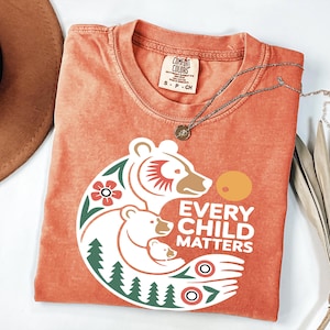 May include: Orange Comfort Colors t-shirt with a graphic of three bears and the text "EVERY CHILD MATTERS". The design includes floral and tree elements. A silver necklace with a pendant is draped across the shirt.