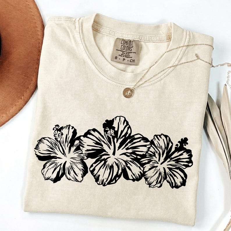 May include: A cream-coloured t-shirt with a black floral design of three hibiscus flowers. The shirt has a crew neck and short sleeves. A gold necklace with a pendant is draped across the shirt. The tag reads "Comfort Colors".