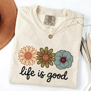 May include: Cream-colored t-shirt with a floral design and the phrase "life is good." The graphic features three flowers in shades of orange, green, and blue. A gold necklace with a pendant is draped across the shirt.