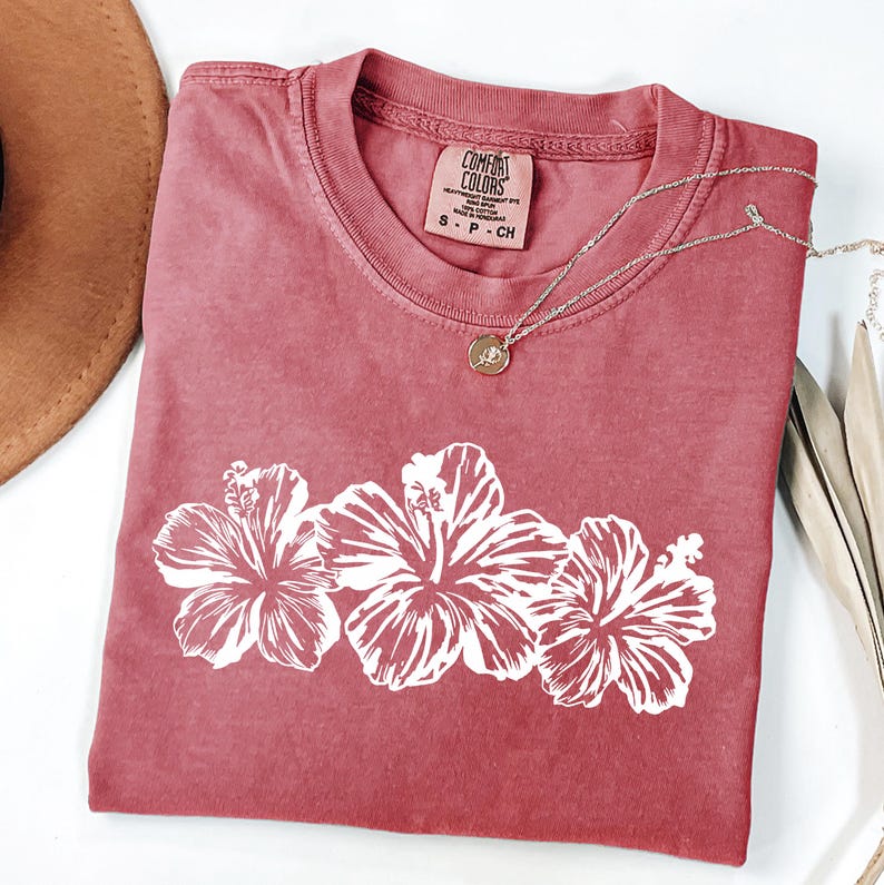 May include: A dusty rose Comfort Colors t-shirt with a white floral design. The shirt features a crew neck and short sleeves. A silver necklace with a pendant rests on the shirt. A brown hat is partially visible in the upper left corner.