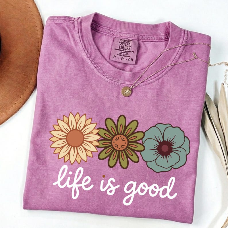 May include: A purple t-shirt with a floral design and the phrase "life is good" in white script. The shirt features three flowers in yellow, green, and blue. A gold necklace with a pendant is draped across the shirt.