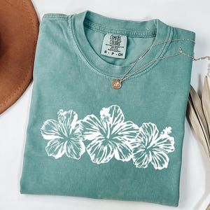 May include: A seafoam green t-shirt featuring a white floral design of three hibiscus flowers. The shirt has a crew neck and short sleeves. A silver necklace with a pendant rests on the shirt. The tag reads "Comfort Colors".