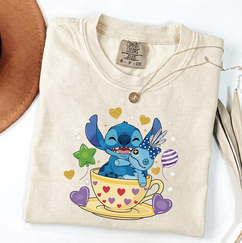 May include: Cream Comfort Colors t-shirt with a Stitch graphic hugging a doll in a teacup. The design includes hearts, stars, and balloons. A gold necklace and a brown hat are also visible. The t-shirt is size S-P-CH.