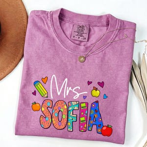 May include: A purple t-shirt with the text "Mrs. SOFIA" in colorful letters and graphics. The design includes a pencil, apples, hearts, and flowers. A gold necklace with a pendant is draped across the shirt.