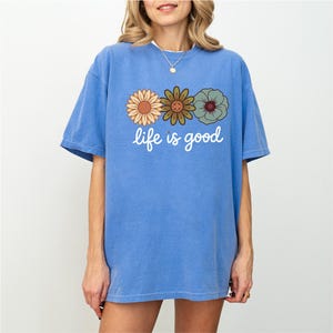 May include: A blue t-shirt with the phrase "life is good" in white script. The shirt features a graphic of three retro-style flowers in shades of orange, green, and blue. The shirt has short sleeves and a relaxed fit.