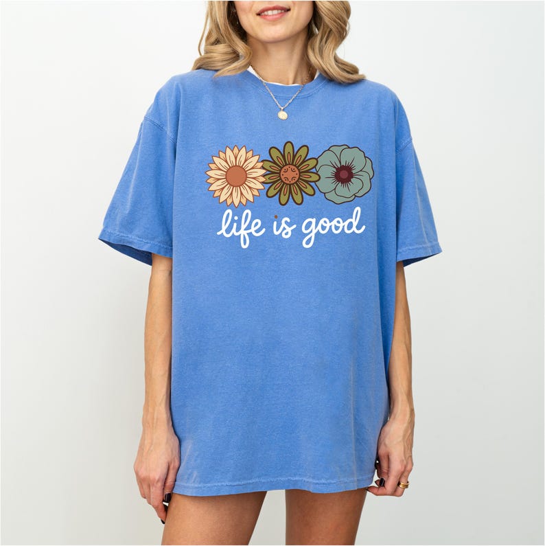 May include: A blue t-shirt with the phrase "life is good" in white script. The shirt features a graphic of three retro-style flowers in shades of orange, green, and blue. The shirt has short sleeves and a relaxed fit.