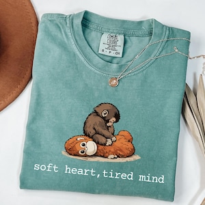 May include: A teal Comfort Colors t-shirt featuring a graphic of two cartoon orangutans with the text "soft heart, tired mind" in white. A silver necklace with a pendant rests on the shirt.