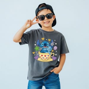 May include: A charcoal grey t-shirt featuring a cartoon image of Stitch and a blue alien character in a yellow teacup with hearts. The shirt is worn by a child wearing sunglasses and a black cap.