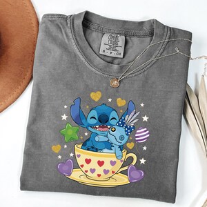 May include: A grey Comfort Colors t-shirt with a graphic of Stitch from Lilo & Stitch hugging a doll in a teacup. The teacup is yellow with red hearts. Gold hearts and stars surround the characters.