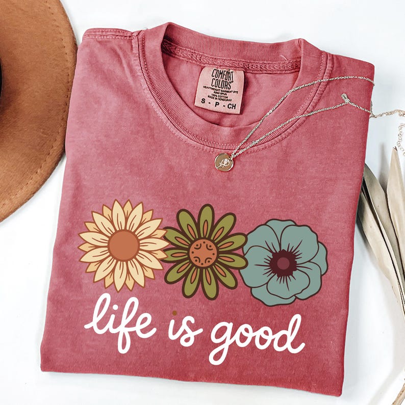 May include: A dusty rose Comfort Colors t-shirt with a floral design and the phrase "life is good" in white script. The graphic features three flowers in yellow, green, and blue. A silver necklace with a pendant is draped across the shirt.
