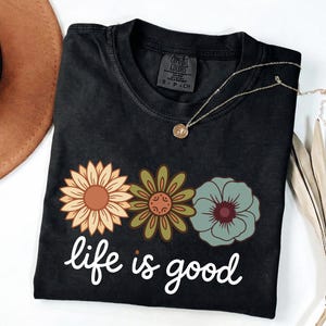 May include: Black t-shirt with a floral design and the phrase "life is good" in white script. The flowers are in shades of orange, green, and blue. A gold necklace with a pendant is draped on the shirt.