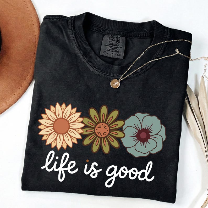 May include: Black t-shirt with a floral design and the phrase "life is good" in white script. The flowers are in shades of orange, green, and blue. A gold necklace with a pendant is draped on the shirt.