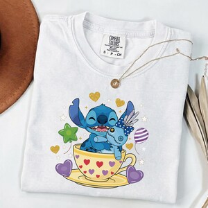 May include: White t-shirt with a cartoon illustration of a blue character hugging a blue doll in a yellow teacup with hearts. The shirt has a Comfort Colors tag and a gold necklace. Gold and purple heart and star graphics.