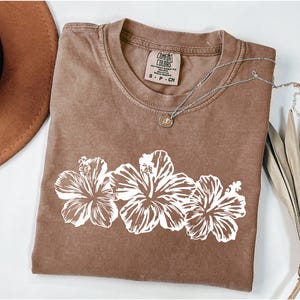 May include: A brown Comfort Colors t-shirt with a white floral design. The shirt has a crew neck and short sleeves. A silver necklace with a pendant is draped across the shirt. The shirt is folded on a white surface.