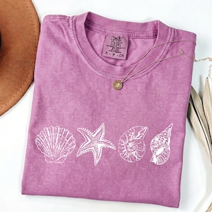 May include: A mauve Comfort Colors t-shirt featuring a white seashell, starfish, and shell design. A gold necklace with a pendant rests on the shirt. The shirt is folded and ready to wear.