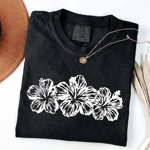 May include: Black Comfort Colors t-shirt with a white floral design of three hibiscus flowers. A gold necklace with a pendant rests on the shirt. A brown hat is visible in the upper left corner.