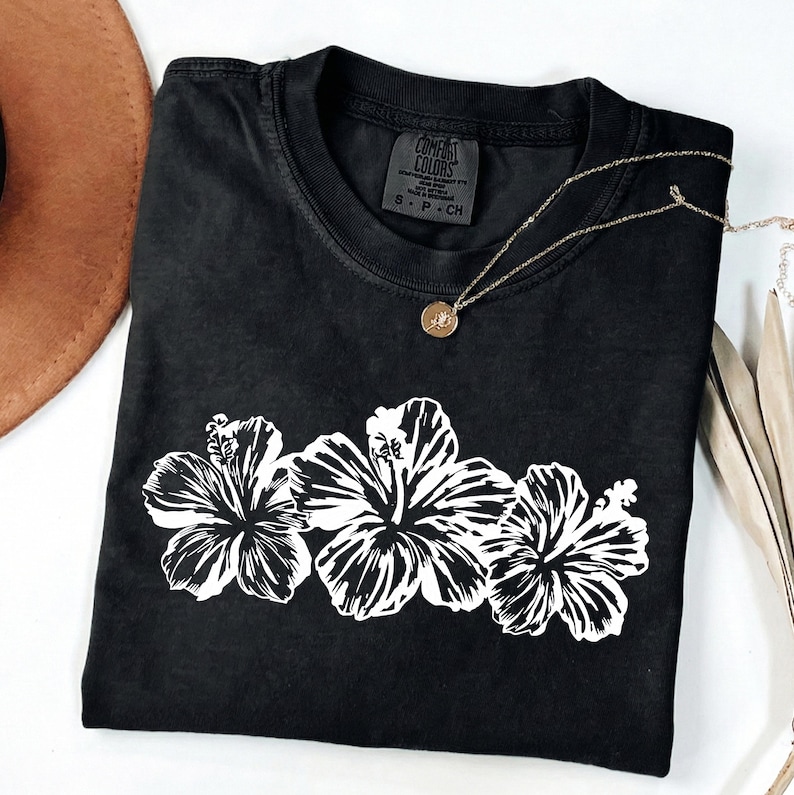 May include: Black Comfort Colors t-shirt with a white floral design of three hibiscus flowers. A gold necklace with a pendant rests on the shirt. A brown hat is visible in the upper left corner.