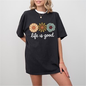 May include: Black t-shirt with the phrase "life is good" in white script. The shirt features three floral designs in shades of yellow, green, and blue. The shirt has short sleeves and a relaxed fit.