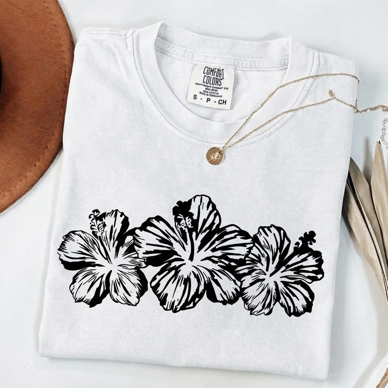 May include: White t-shirt featuring a black hibiscus flower design. The shirt has a crew neck and short sleeves. A gold necklace with a pendant is draped across the shirt. The tag reads "Comfort Colors".