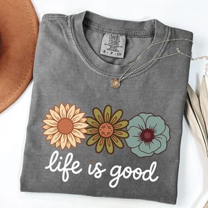 May include: A gray t-shirt with a floral design and the phrase "life is good" in white script. The shirt features three flowers in shades of orange, green, and blue. A silver necklace with a small pendant is draped across the shirt.